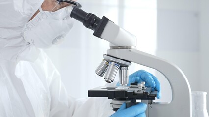 Medical virologist in a hazmat suit, mask, and blue gloves, diligently working at a microscope in a...