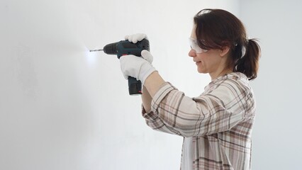 Middle aged female professional worker wearing protective gear and gloves, is drilling hole in...