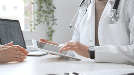 Doctor man in lab coat and stethoscope listening to patient while using tablet in modern medical...