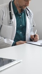 Doctor man wearing lab coat over green shirt, and stethoscope is writing medical prescription or...