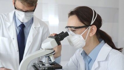 Two scientists in lab coats and safety gear conducting research, one observing through a microscope...
