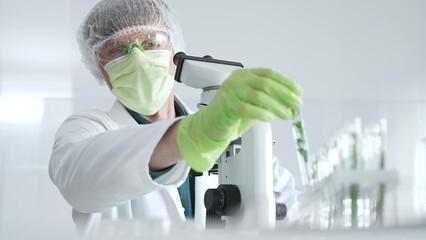 Adult male scientist wearing green protective gear looking at plant samples in test tubes and using...