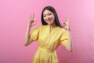 Asian woman in a yellow dress smiling and making a double OK hand gesture with both hands against a...