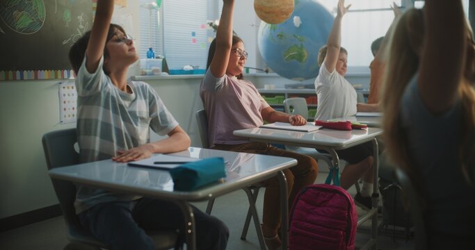 Elementary School Students Sitting at the Desks, Listening to Lecture from Teacher, Raising Hands to Provide Correct Answer. Team of Smart Diverse Kids Studying Science, Geography in Modern Classroom.