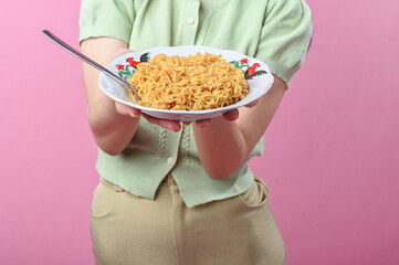 Asian woman smiling while holding a plate of cooked instant noodles with a spoon, wearing a green...