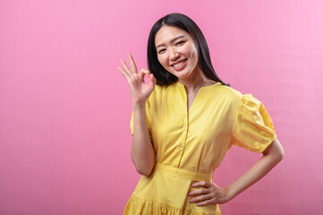 Asian woman wearing a bright yellow dress smiling and showing a peace sign with one hand while the...