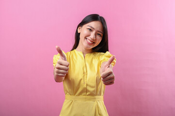 Asian woman in a yellow dress smiling with both thumbs up against a bright pink background....