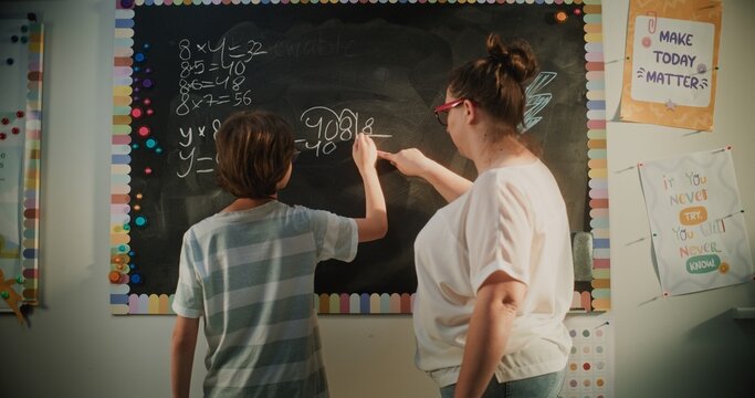 Smart Pupil Showcasing Math Knowledge: Static Shot of Elementary School Boy Solving Mathematical Problems Writing on the Chalkboard in Classroom. Female Teacher Checking Answers, Helping Student.