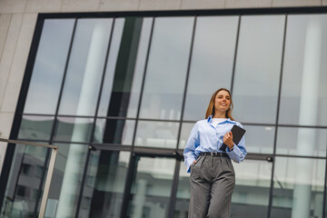 A confident businesswoman walks outside a modern office building, holding a tablet, showcasing her...