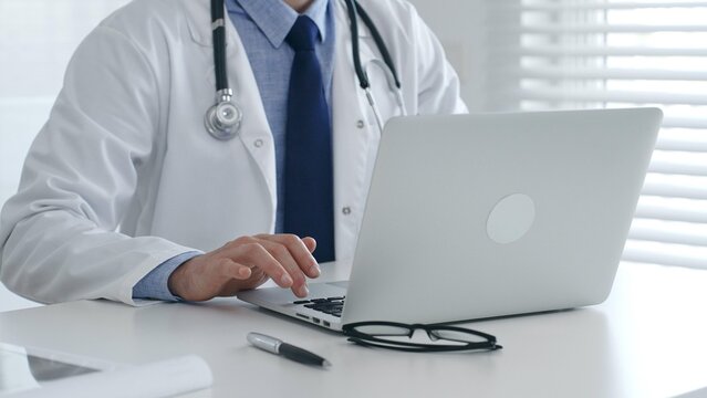 Medical doctor wearing lab coat and stethoscope is sitting at desk and typing on laptop, with eyeglasses and pen nearby, in bright medical office. Medicine and health care concept