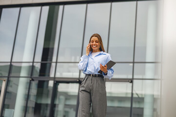 A cheerful businesswoman engaged in a phone call, expertly holding a tablet, dressed in stylish...