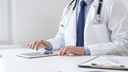 Doctor man wearing white medical coat and stethoscope using digital tablet while sitting at desk in medical office, with medical chart and pen in the foreground. Medicine concept