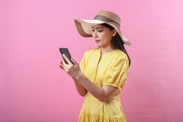 Asian woman in yellow dress and summer hat using smartphone against a pink background in studio...