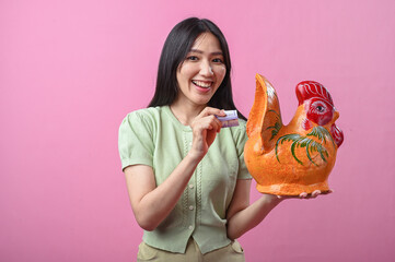 Asian woman smiling while holding a ceramic rooster and inserting a banknote, suggesting savings or...