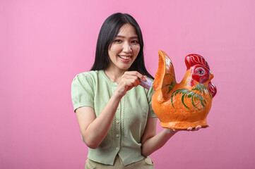 Asian woman smiling while holding a ceramic rooster and inserting a banknote, suggesting savings or...