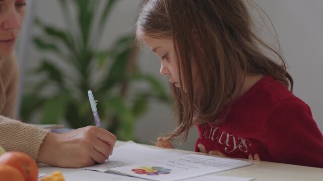 Mother and daughter sitting at home table doing homework together. Child focused on writing while parent gently guides her with support