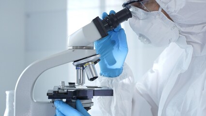 Medical virologist in a hazmat suit, mask, and gloves, diligently working at a microscope in a...