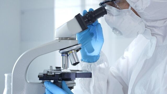 Medical virologist in a hazmat suit, mask, and gloves, diligently working at a microscope in a laboratory researching viruses. Healthcare and science concept