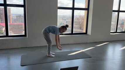 A young child stretches while practicing an online workout in a bright room with large windows.