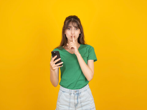 Hush it's a secret. Portrait of young caucasian brunette 20s woman hold use phone in hand put finger on lips looking camera serious face expression wear casual green t-shirt isolated on yellow.