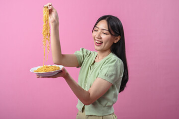 Asian woman in green top smiling while lifting a forkful of cooked instant noodles from a plate,...