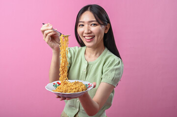 Asian woman in green top smiling while lifting a forkful of cooked instant noodles from a plate,...