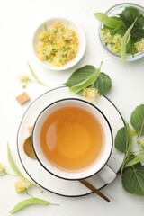 Tasty linden tea in cup, spoon, sugar cubes, leaves and flowers on white table, flat lay