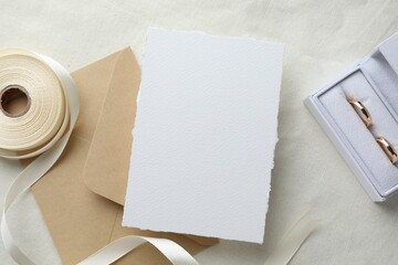 Wedding invitation card, envelope, golden rings and ribbon on white cloth, flat lay
