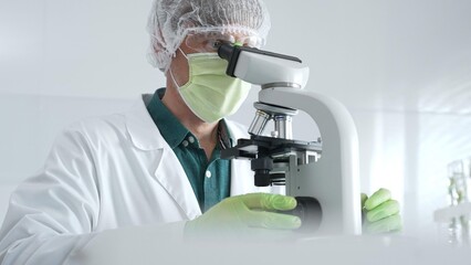 Medical scientist wearing protective mask, cap and green gloves using microscope doing scientific...
