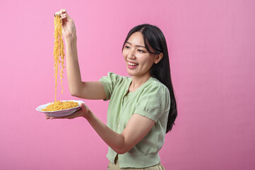 Asian woman in green top smiling while lifting a forkful of cooked instant noodles from a plate,...