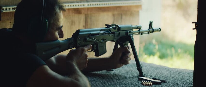 Man with earmuffs fires rifle with visible muzzle flash while prone on mat during outdoor training