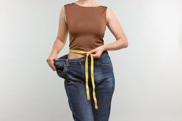 Woman with measuring tape wearing old jeans after weight loss on light grey background, closeup