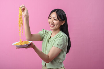 Asian woman in green top smiling while lifting a forkful of cooked instant noodles from a plate,...