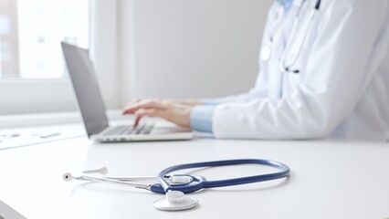 Closeup of blue stethoscope is lying on the table near female doctor working on laptop,...