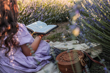 Girl in lavender dress reading a Bible in blooming lavender field. Peaceful summer moment with...