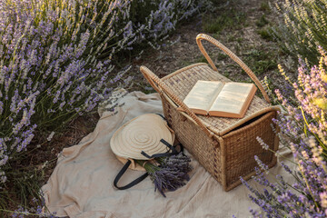 Wicker picnic basket with open book, lavender bouquet, and straw hat on light fabric in blooming...
