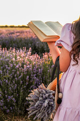 Girl in lavender field reading book at sunset. Holding a straw bag with lavender bouquet, dressed...