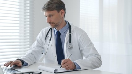 A male doctor in a lab coat with a stethoscope sits at a table in a bright medical office and uses...