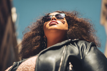 Confident African-American woman in sunglasses and leather jacket viewed from below, strong urban vibe with bright sky, perfect for fashion and streetwear themes. female, trendy, independence