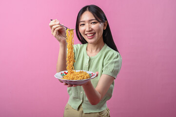 Asian woman in green top smiling while lifting a forkful of cooked instant noodles from a plate,...
