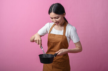 Asian woman in brown apron stirring ingredients in a black saucepan with a whisk, smiling in front...