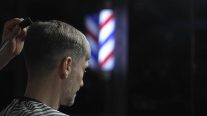 Professional barber grooming male client's hair with scissors, vintage barber pole framing...