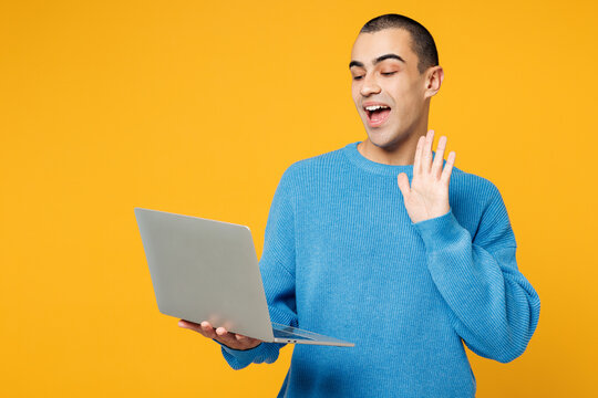 Young IT middle eastern man he wearing blue sweatshirt casual clothes hold use work on laptop pc computer waving hand talk speak isolated on plain yellow orange background studio. Lifestyle concept.