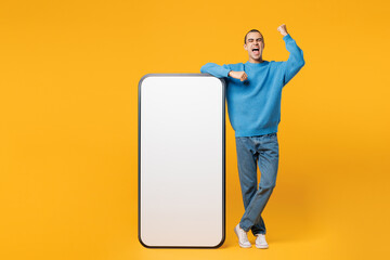 Full body young man wears blue sweatshirt casual clothes big huge blank screen mobile cell phone smartphone with area do winner gesture isolated on plain yellow orange background. Lifestyle concept.