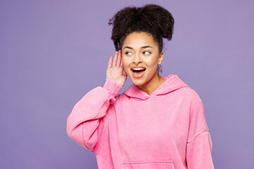 Young curious nosy happy woman of African American ethnicity wear pink hoodie casual clothes try to hear you overhear listening intently isolated on plain pastel purple background. Lifestyle concept.
