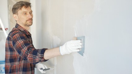Male construction worker spreading plaster on white wall, wearing protective gloves during home...