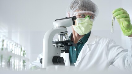 Adult male scientist wearing green protective gear looking at plant samples in test tubes and using...