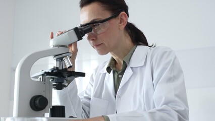 Female scientist in a lab coat and protective glasses examining samples through a microscope in a...