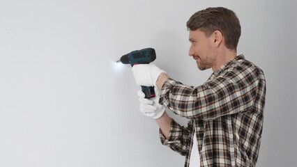 Male construction worker in checked shirt and safety gloves is screwing a screw into the wall using...