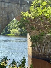 Old stone bridge arches over a calm river surrounded by lush greenery and sunlit foliage. A...